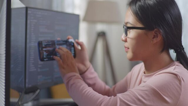 Close Up Of Asian Teen Girl Programmer Using Smartphone While Creating Software Engineer Developing App, Program, Video Game On Destop Computer At Home
