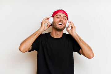 Indoor shot of handsome modern European guy in red headband listens music keeps arms on headphones has overjoyed expression looks at the camera poses in black casual t-shirt over white background
