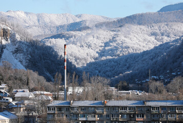 Beautiful winter cityscape in the foothills. The pipe of a gas boiler house on the background of snow-capped mountains. Sochi, Russia.