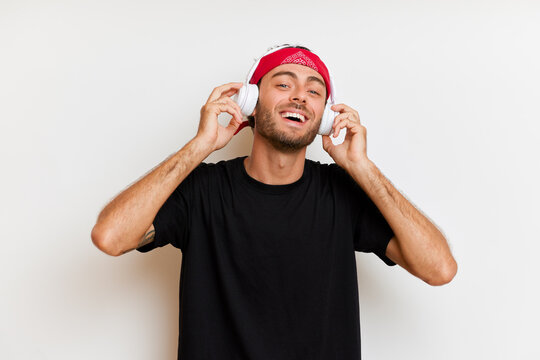 Indoor shot of handsome modern European guy in red headband listens music keeps arms on headphones has overjoyed expression looks at the camera poses in black casual t-shirt over white background