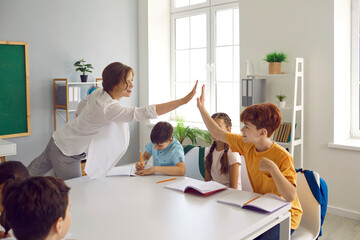 Teacher and student give high fives to each other after successfully completing task in class. Friendly cheerful female teacher encouraging her little students in school classroom. Education concept.