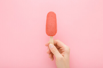 Young adult woman hand holding red strawberry ice cream with stick on light pink table background....