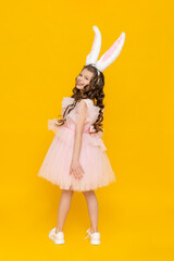 Easter children's holiday. A teenage girl in a lush dress rejoices in spring. The child is wearing long rabbit ears. A beautiful little girl with long hair, full-length on a yellow isolated background