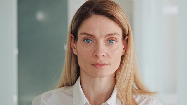 Head Shot Close Up Portrait Of Serious Focused Millennial Dreamy Successful Business Woman Looking At Camera. Female Leader Have Natural Makeup, Blue Eyes Dreseed In White Shirt Standing In Office.