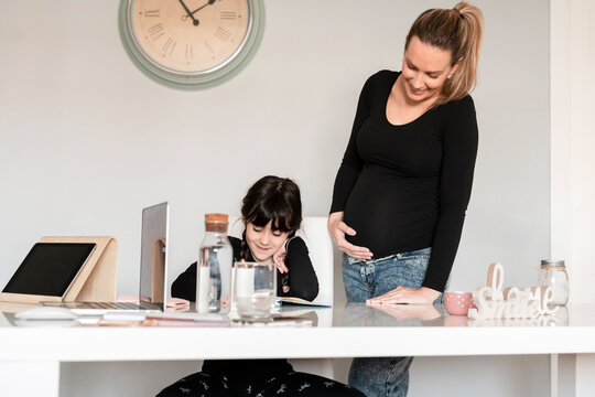 Pregnant mother and toddler daughter using laptop at dining table