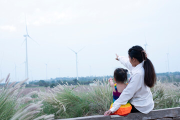 Beautiful young mother daughter relax sitting summer meadow background and windmill,Wind turbines are alternative electricity sources.