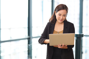 Portrait of confident young asian businesswoman researching and planning work with happy smiling face while using computer at business meeting in modern office building and city background