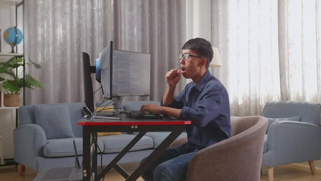 Asian Boy Programmer Yawning While Creating Innovative Software Engineer Developing App, Program, Video Game On Desktop Computer At Home. Terminal With Coding Language 
