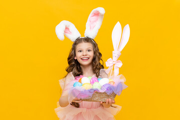 Obraz na płótnie Canvas Easter for children. A little girl holds a basket with colorful eggs for games on an Easter spring day. Charming teenage girl with long rabbit ears on a yellow isolated background.