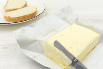 Fresh piece of butter on the packaging paper, knife and a plate of bread.