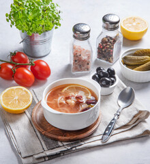 Solyanka soup is a traditional Russian soup in a bowl with olives, meat and pickles on a bright table with fresh tomatoes, lemon and herbs.