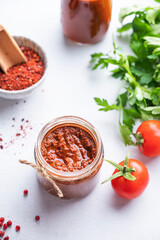 Hot sauce adjika. Homemade appetizer with pepper and tomatoes in a jar on light background with fresh herb and vegetables. Top view.