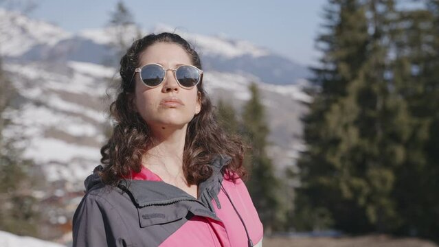 Young woman in colorful winter coat and sunglasses looking at the snowy view. Female skier in ski resort on winter vacation holiday enjoying view of mountains and trees on snowy slopes on a sunny day