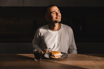 Dreaming man sitting near table look at side in kitchen at home touches his stomach, burger on the table. Happy smiling man think about fast food, unhealthy food.