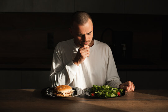 Man make decision between salad and burger. Man think and make choice between health food vs fast food. Healthy nutrition, dieting, eating disorder. Modern kitchen interior. Man look at salad.