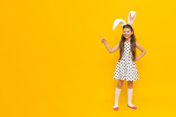 A cheerful girl with rabbit ears on her head and chicken eggs in her hands on a yellow background. pointing to the side of the advertisement, a funny happy child. Easter child. Easter holiday.