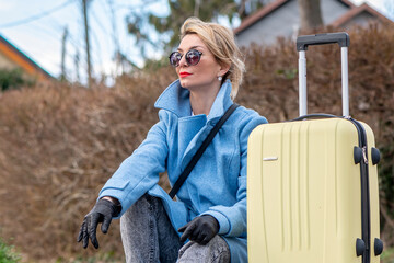 a 40-45 blonde woman with glasses and a suitcase is sitting on a blurry background of houses and...