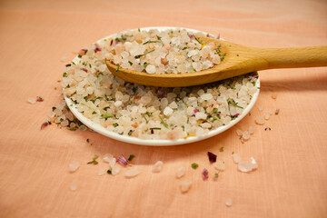 Salt with herbals on a white plate, a wooden spoon with salt on it lying on this plate, orange - pink background, close up