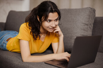 Fototapeta premium Happy indian or arabian young woman lying sofa and using laptop. Remote working