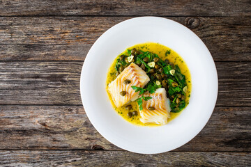 Fish dish - fried cod with spinach and capers in saffron sauce on wooden table

