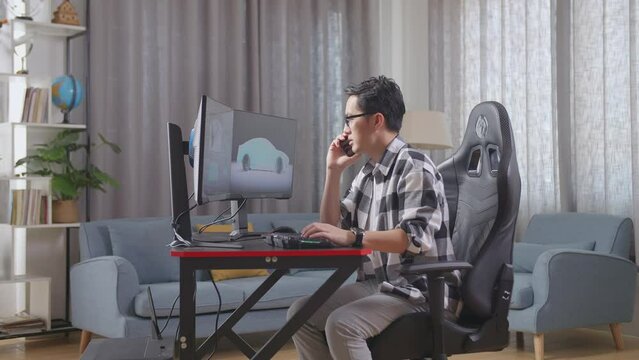 Side View Of Asian Male Automotive Designer Talking On Smartphone While Working On 3D Model Of Ev Car On The Desktop Computers At Home
