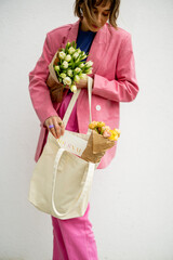 Stylish woman in pink suit takes some magazine from eco handbag and holds flowers on white background. Canvas bag with blank space