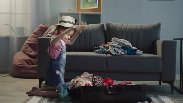 Pretty preschooler girl with hat puts clothes pile into suitcase and imitates airplane at home. Happy child prepares for first flight abroad