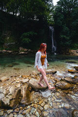 Beautiful girl with red hair enjoying life and beautiful nature. Behind the Abhesi Waterfall, Stones in the mountain river, Kutaisi, Georgia. Vertical photo.