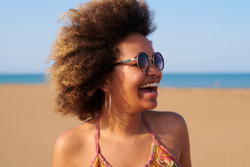 Emotional woman with frizzy hair and brackets. Portrait of laughing pretty woman dyed afro hairstyle, sunglasses, brackets and big hoop earrings on the beach against unfocused sea in background.