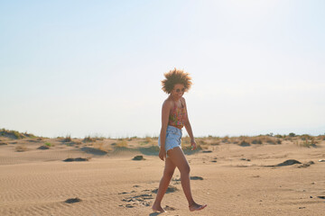 Laughing woman walking on sand. Full length portrait of attractive smiling woman in sunglasses with frizzy hair walking on sand in sunlight.