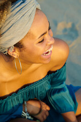 Stylish woman smiling in sunlight. Above portrait of attractive happy woman in band, earrings and dress with bare shoulders laughing sitting on sand in sunlight.