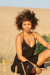 Beautiful ethnic woman sitting on beach. Young beautiful black woman with Afro hairstyle wearing stylish beach dress and looking at camera sitting on sand
