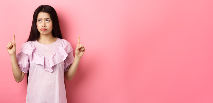 Sad Pitiful Asian Girl Sulking, Pointing And Looking Up With Disappointed Face, Feeling Upset, Standing Against Pink Background