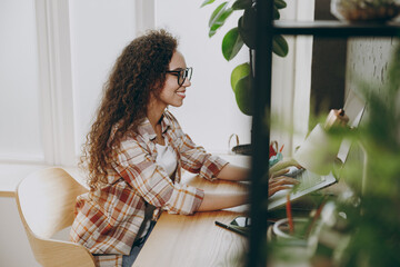 Side profile view young smiling happy smart successful employee IT business woman of African American ethnicity wear casual shirt glasses sitting work at office desk with laptop pc computer indoors.