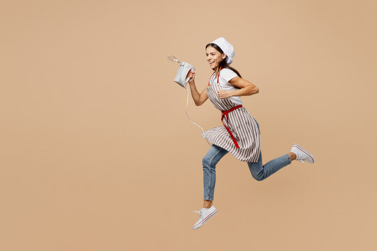 Full body sideways happy smiling young housewife housekeeper chef baker latin woman wear apron toque hat jump high hold in hand mixer isolated on plain pastel light beige background Cook food concept