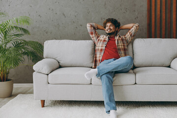 Young fun Indian man wearing casual clothes hold hands behind neck looking aside sits on grey sofa couch stay at home hotel flat rest relax spend free spare time in living room indoor. Lounge concept.