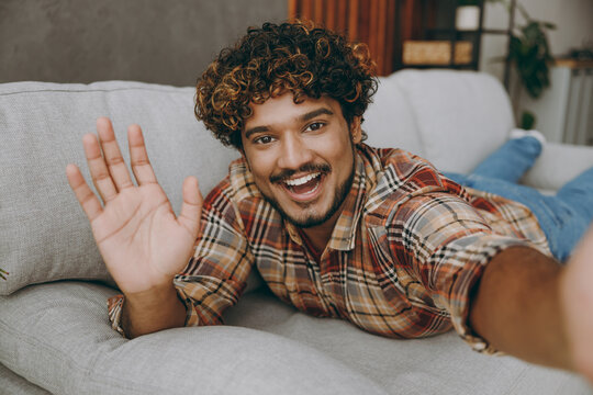 Close up young Indian man wear casual clothes do selfie shot pov on mobile cell phone waving hand lay down on grey sofa couch stay at home hotel flat rest relax spend free spare time in living room