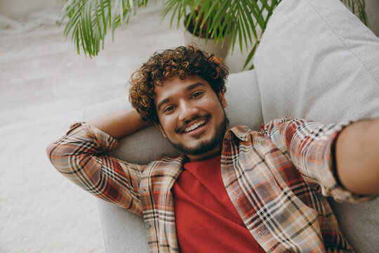 Close up top view young Indian man wear casual clothes doing selfie shot pov on mobile cell phone lay down on grey sofa couch stay at home hotel flat rest relax spend free spare time in living room.