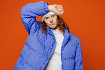Young tired sick sad ill man with long curly hair wears hat purple ski padded jacket casual clothes put hand on forehead suffer from headache isolated on plain orange red background studio portrait. © ViDi Studio
