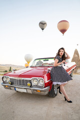 beautiful girl in retro style posing near a vintage red cabriolet car on background of balloons in...