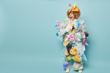 Horizontal shot of displeased annoyed woman covered with plastic garbage collects litter to clean nature from pollution points index finger aside on blank space isolated over blue background.