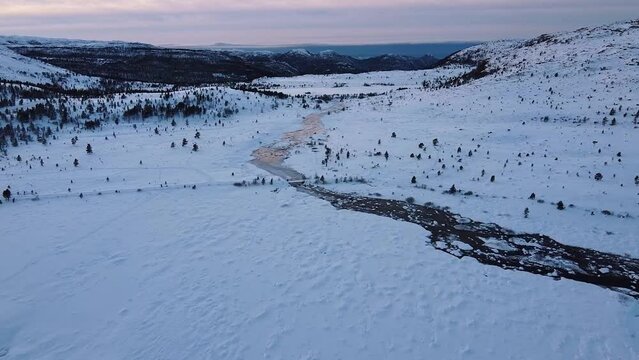 Aerial view of snow and an icy river in the snowy mountains in the picturesque Norwegian nature during a pink sunset. A panoramic view from a drone of a popular cross country ski trail in Norway. 