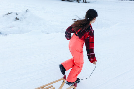 Girl In Red Pullover And Orange Ski Pants Runs Away With Wooden Sled On Snowy Road