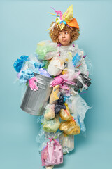 Vertical shot of unhappy curly haired woman volunteer holds rubbish bin covered with plastic garbage looks upset aside because of environmental pollution manages wastes isolated over blue background