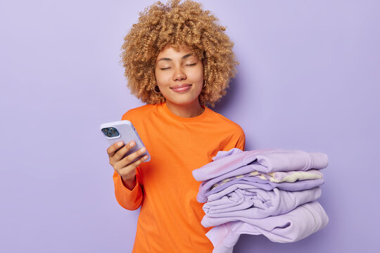 Horizontal shot of pleased curly haired woman wears casual orange jumper holds mobile phone and folded laundry keeps eyes closed from satisfaction isolated over purple background does housework - Powered by Adobe