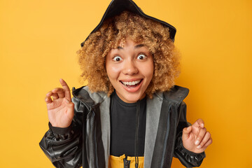 Happy surprised woman feels very glad stares impressed at camera smiles broadly shows white teeth wears hat and black leather raincoat cannot believe in something awesome isolated on yellow background