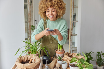 Positive woman takes photo of potted plants with smartphone demonstrates her results of...