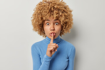 Fototapeta na wymiar Surprised mysterious young woman with curly hair presses index finger to lips makes silence gesture dressed in casual blue turtleneck isolated over white background has secret. Be quiet sign.