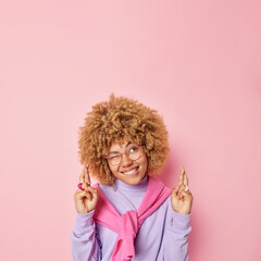 Studio shot of cheerful hopeful woman bites lips winks eye crosses fingers anticipaes good news yearns and waits for something dressed casually isolated over pink background copy space for promotion