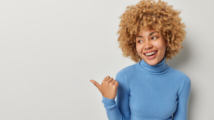 Smiling curly haired woman points thumb aside shows advertisement or promo offer wears casual blue...